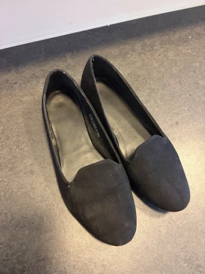 Svarta ballerinaskor i mockaimitation - Säljer ett par svarta ballerinaskor i mockaimitation med klassisk, stilren design och rundad tå. Skorna har en låg klack och enkel slip-in-modell som passar till många olika outfits.