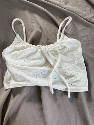 Vit broderad linnetopp med knyt - Söt vit linnetopp med broderade detaljer och knyt framtill. Toppen är ärmlös med smala axelband och har en croppad passform. Perfekt för varma dagar och enkel att styla till olika outfits.