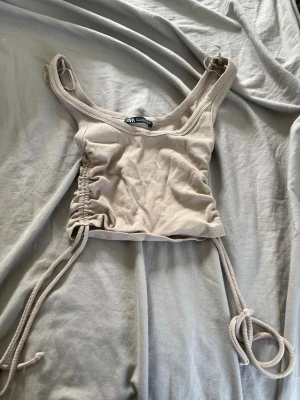 Beige ribbad croppad topp från Zara - Trendig beige croppad topp från Zara med ribbad struktur och snörning i sidorna. Toppen är ärmlös och har breda axelband, perfekt för en avslappnad och cool stil.