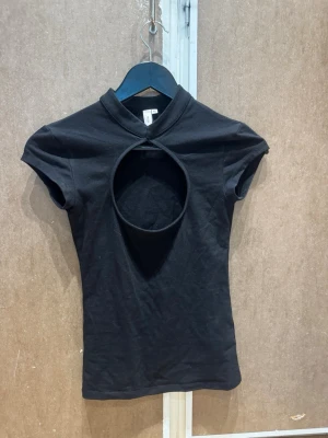 Svart topp med cut out-detalj - Säljer en svart kortärmad topp med hög krage och stor rund cut out-detalj framtill. Toppen har en slim passform och är tillverkad i mjukt bomullsmaterial. Perfekt för dig som vill ha en edgy och trendig look. Den är aldrig använd🥰
