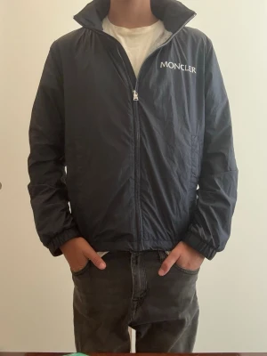 Svart vindjacka från Moncler - Snygg svart vindjacka från Moncler med dragkedja framtill och hög krage. Jackan har elastiska muddar vid ärmsluten och logotyp på bröstet samt ärmen. Tillverkad i lätt och tunt material som passar perfekt till blåsiga dagar. Ny pris ungefär 6 tusen säljer för 1500