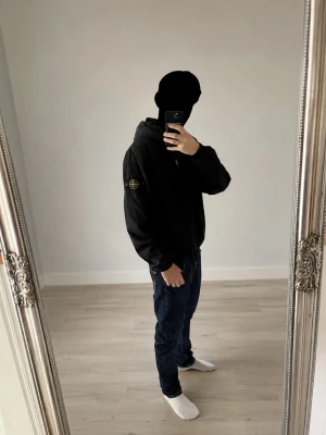 Svart Stone Island vindjacka med huva - Säljer en svart vindjacka från Stone Island med huva och dragkedja framtill. Jackan har den klassiska logotypen på vänster ärm och justerbar nederkant. Perfekt för dig som vill ha en stilren och funktionell jacka till vardags.