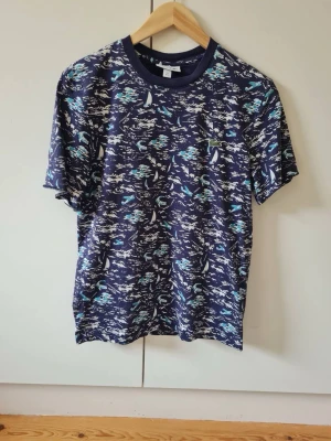 Mönstrad marinblå t-shirt från Lacoste strl S - Marinblå t-shirt från Lacoste med mönster i vitt och turkost. T-shirten har rund hals och korta ärmar. Material 100% bomull. Storlek S.