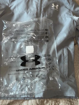 Ljusgrå vindjacka från Under Armour - Snygg ljusgrå vindjacka från Under Armour med dragkedja framtill och huva. Jackan har två sidofickor och logga på bröstet. Tillverkad i lätt och vindtätt material, perfekt för blåsiga dagar. Enkel och stilren design som passar till det mesta.