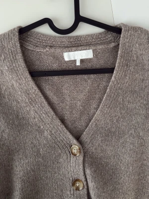 Beige kofta med knappar från NA-KD - Mysig beige kofta från NA-KD med v-ringning och 4 knappar framtill. Koftan är stickad i ett mjukt material och har långa ärmar samt ribbade muddar vid ärmslut och nederkant. Perfekt för lager på lager och enkel att matcha.