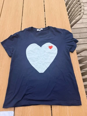 Mörkblå t-shirt med stort hjärta - Snygg mörkblå t-shirt med ett stort vitt hjärta på bröstet och en liten röd hjärtdetalj. Klassisk rund hals och korta ärmar. Perfekt för dig som vill sticka ut med en enkel men cool design.