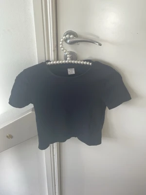 Svart croppad ribbad t-shirt - Säljer en svart croppad t-shirt med ribbad struktur. T-shirten har korta ärmar och rund halsringning. Perfekt till jeans eller kjol för en enkel och stilren look.
