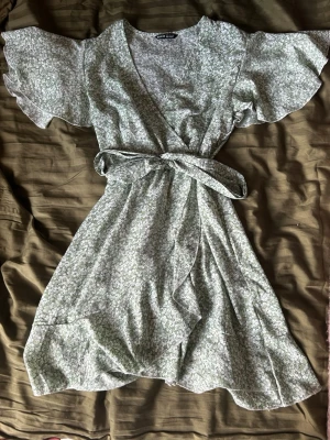 Grön blommig omlottklänning SHEIN - Söt grön omlottklänning från SHEIN med vitt blommönster. Klänningen har korta, vida ärmar och knytskärp i midjan. Lätt och luftig modell som passar perfekt till sommaren. Det finns en dragkedja på sidan som gör den enklare att få på.