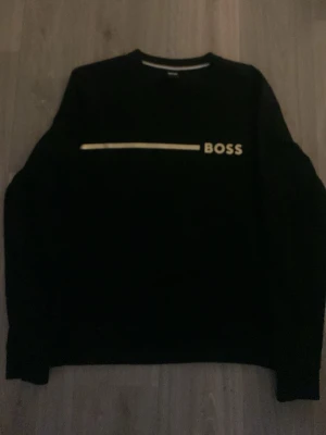 Svart stickad tröja med BOSS-tryck - Svart stickad tröja med rund hals och lång ärm. På bröstet finns ett vitt BOSS-tryck och en vit linje. Tröjan har en klassisk passform och ribbade muddar vid ärmslut och nederkant. Perfekt för dig som gillar stilrena och enkla plagg.