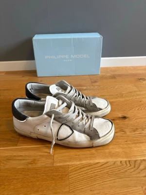Vita Philippe Model sneakers med blå mocka - Snygga vita sneakers från Philippe Model med grå detaljer och blå mocka häl. Skorna har en klassisk låg siluett, vita snören och en robust sula. Tillverkade i läder med mockadetaljer för en trendig look. 5/10 skick då dom är rätt använda men kan fixas av lite städning jag fixar innan köp! Sulorna har låsnat lite på båda skorna. 