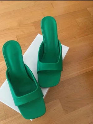 Gröna pumps med fyrkantig tå - Snygga gröna pumps med öppen häl och bred rem över foten. Skorna har en fyrkantig tå och en stabil klack, vilket ger en modern och trendig look. Perfekta för dig som vill sticka ut med färg och stil. Materialet ser ut att vara konstläder.Endast använd en gång.