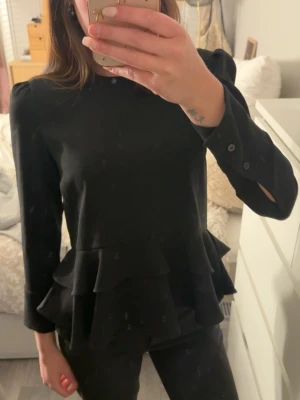 Svart volangblus med långa ärmar - Snygg svart blus med volangdetaljer i midjan och långa ärmar med knapp vid ärmslutet. Blusen har en rak passform och rund halsringning, perfekt för dig som gillar stilrena plagg med en twist.