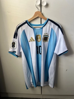  Messi matchtröja - Säljer en officiell Argentina matchtröja med Messi och nummer 10 på ryggen. Tröjan har klassiska blå och vita ränder, guldiga detaljer och FIFA-märke på bröstet. Korta ärmar och rund hals, tillverkad i lätt och ventilerande material. Perfekt för fotbollsfans!