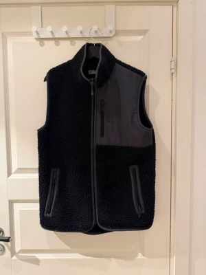 Svart fleeceväst med dragkedjor - Säljer en svart fleeceväst med hög krage och hel dragkedja framtill. Västen har två sidofickor och en bröstficka med dragkedja samt en snygg detalj i grått tyg över bröstet. Perfekt lager på lager och riktigt skön att bära.