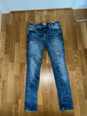 Blå slitna jeans från Jack & Jones - Säljer ett par blå jeans från Jack & Jones med slitna detaljer och klassisk femficksdesign. Jeansen har en slim passform och är tillverkade i bomull. Perfekta för en avslappnad och trendig look.
