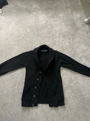Stickad Cardigan  - Stilren svart stickad cardigan från Jack&Jones med snygg krage och knappar framtill. Två praktiska fickor på framsidorna. Perfekt att slänga på sig över en t-shirt för snygg och stilren look. Pris kan såklart diskuteras!