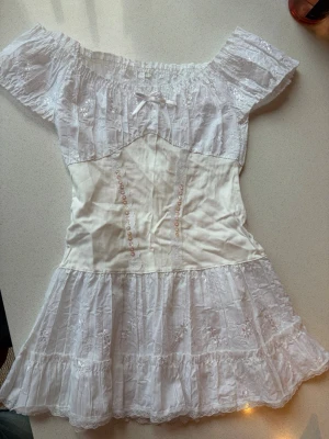 Vit offshoulder kortklänning med spets - Söt vit kortklänning med offshoulder och volangdetaljer. Klänningen har spets överdel, liten rosett framtill och broderade detaljer. Kjolen är volangig och har ett romantiskt uttryck. Perfekt för sommarens alla tillfällen.