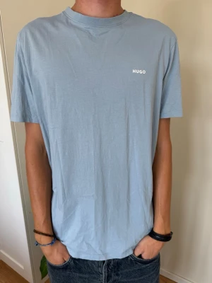 Ljusblå t-shirt från HUGO - Snygg ljusblå t-shirt från HUGO med minimalistisk logga på bröstet. Klassisk passform med rund hals och korta ärmar. Tillverkad i mjuk bomull som känns skön mot huden. Perfekt till jeans eller shorts för en clean look.