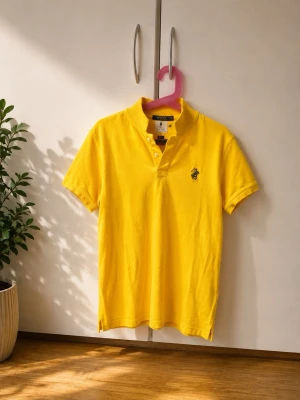 Gul pikétröja från Polo Ralph Lauren - Klassisk gul pikétröja från Polo Ralph Lauren med korta ärmar och knappslå framtill. Tröjan har den ikoniska broderade loggan på bröstet och är tillverkad i mjuk bomull. Perfekt för en avslappnad och stilren look.