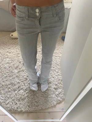 Ljusgrå lågmidjade bootcut jeans med dubbla knappar - Säljer ett par ljusgrå jeans med bootcut-modell och dubbla knappar i midjan. Jeansen har klassiska fem fickor och är i ett mjukt denimtyg. Perfekta för dig som gillar en avslappnad men ändå trendig look.