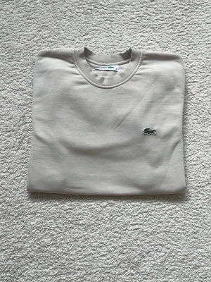 Beige sweatshirt från Lacoste - Säljer en stilren beige sweatshirt från Lacoste med den klassiska gröna krokodilloggan broderad på bröstet. Tröjan har rund halsringning, långa ärmar och ribbade muddar vid ärmslut och nederkant. Perfekt för en clean och avslappnad look.