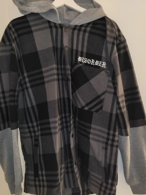 Gråsvart rutig overshirt med luva - Säljer en oversized overshirt i grått och svart rutigt mönster med trycket 'DISORDER' på bröstfickan. Jackan har gråmelerade sweatshirt-ärmar och luva, samt knappar framtill. Perfekt för en avslappnad streetwear-look.