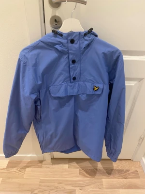 Blå lyle scott vindjacka med huva - Snygg blå vindjacka från lyle scott med huva och knappstängning framtill. Jackan har en gul logga på bröstet och elastiska muddar vid ärmsluten. Perfekt för blåsiga dagar och enkel att matcha till olika outfits.