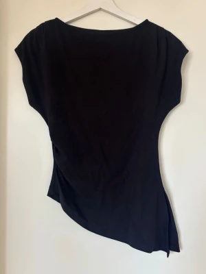 Svart asymmetrisk topp med slits - Säljer en svart topp med asymmetrisk form och slits i sidan. Toppen har korta ärmar och är tillverkad i mjukt material som känns skönt mot huden. Perfekt för dig som gillar en stilren men edgy look.