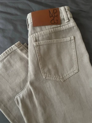 Beige vida jeans med - Säljer ett par beige jeans med breda ben och hög midja. Jeansen har två stora bakfickor och en läderpatch med tryck i midjan. Materialet är kraftig denim i bomull och passformen är avslappnad och trendig.