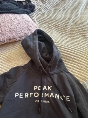 Mörkgrå hoodie Peak Performance - Snygg mörkgrå hoodie från Peak Performance med vit logga framtill. Klassisk modell med huva och dragsko, samt mjukt material som känns skönt mot huden. Perfekt för en avslappnad och sportig stil.