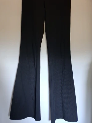 Svarta ribbade utsvängda leggings - Säljer ett par svarta leggings med ribbad struktur och utsvängda ben. Materialet är stretchigt och följsamt, perfekt för en trendig och bekväm look. Diskreta tunna ränder i tyget ger extra detalj. Passar dig som gillar stilrena och moderna plagg.