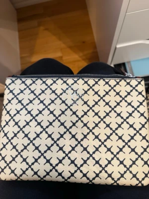Beige kuvertväska med svart mönster - Snygg beige kuvertväska med svart grafiskt mönster och silverfärgad dragkedja. Väskan har en stilren och modern look med ett diskret emblem framtill. Tillverkad i ett tåligt material som känns lyxigt.