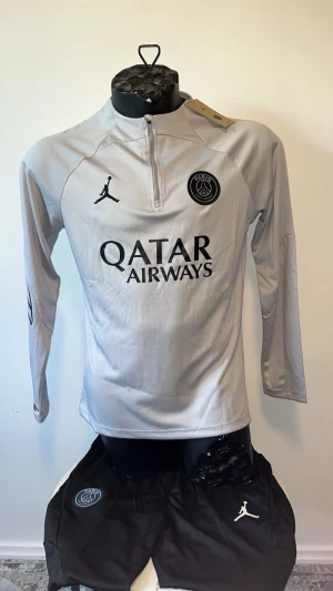 PSG träningsset grå och svart - Snyggt träningsset från PSG med långärmad grå tröja och svarta byxor. Tröjan har halv dragkedja, PSG-logga och Jordan-logga samt Qatar Airways-tryck. Byxorna har PSG-märke, Jordan-logga och Paris-tryck. Perfekt för fotbollsträning eller chill.