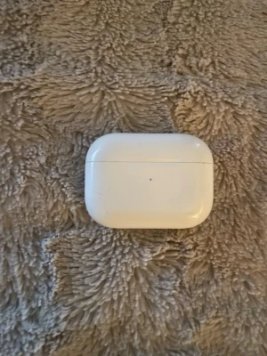 AirPod Pro 1 - Säljer ett par vita trådlösa in-ear hörlurar med laddningsetui. Etuiet är kompakt och har rundade kanter, perfekt att ta med i fickan eller väskan. Stilren design i blank plast som ger en modern känsla.