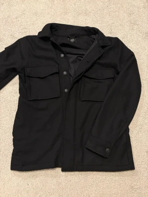 Svart overshirt från Neuw - Svar overshirt ifrån NEUW. Overshirten har praktiska fickor både på bröstet (se bild 4) samt sidofickor (se bild 5).🙌  Materialet består av 50 % ull och 50 % polyester, vilket gör plagget extra tjockt, varmt och av hög kvalitet, perfekt under denna tiden på året🤩  Hör av dig vid minsta fundering kring mått eller passformsbilder😁