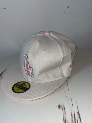 Vit New Era LA keps med rosa detaljer - Snygg vit keps från New Era med LA-logga i ljusrosa framtill och rosa knapp på toppen. Kepsen har broderad detalj på sidan och klassisk 59FIFTY-form med rak skärm. Perfekt för dig som vill sticka ut med stilrena färger och coola broderier.