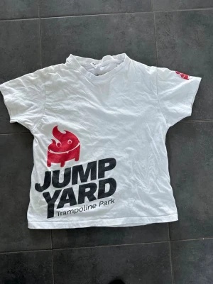 Vit Jump Yard t-shirt med tryck - Vit t-shirt med stort svart och rött Jump Yard Trampoline Park-tryck framtill och en liten röd detalj på ärmen. Klassisk passform och rund hals. Perfekt för dig som gillar sportiga och lekfulla plagg.