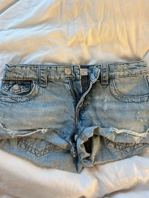 Jeansshorts - Säljer ett par ljusblå jeansshorts med slitna detaljer och råa kanter. Shortsen har broderade bakfickor med snyggt mönster och knappar både fram och bak. Perfekta för sommaren och ger en avslappnad vibe.