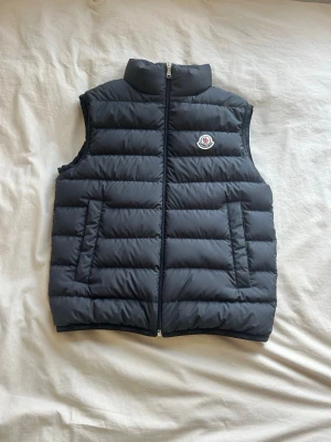 Svart dunväst från Moncler - (Pris kan diskuteras )Snygg svart dunväst från Moncler med quiltad design och hög krage. Västen har dragkedja framtill, två fickor och Moncler-logga på bröstet. Perfekt för lager-på-lager och håller dig varm med stil.