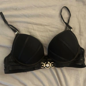 Svart push-up bh med gulddetalj - Snygg svart push-up bh med vadderade kupor och justerbara axelband. Framtill sitter en dekorativ guldfärgad detalj med strass. Bh:n har meshpartier på sidorna och stängs med hyskor bak. Perfekt för dig som vill ha en elegant och stilren look.