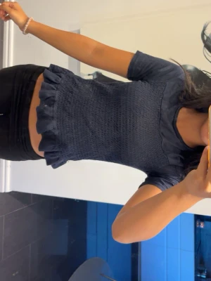 Mörkblå topp med volangkant - Säljer en mörkblå topp med korta ärmar och smockad midja. Toppen har en söt volangkant nedtill och är croppad i modellen. Perfekt för dig som gillar en feminin och trendig stil.
