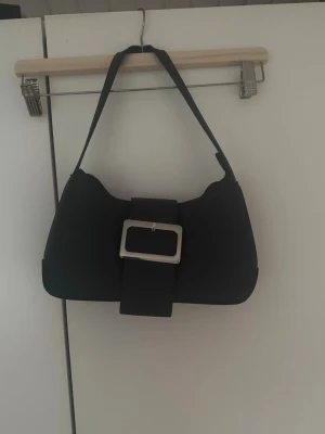 Svart axelremsväska med stor spänne - Snygg svart handväska i axelremsmodell med en stor, silverfärgad spännesdetalj framtill. Väskan har en mjuk, rundad form och dragkedja baktill för extra förvaring. Materialet ser ut att vara konstläder. Perfekt för dig som gillar minimalistisk och trendig stil. Bandet har en liten böjd del men det är inget som märks när väskan andvändes 