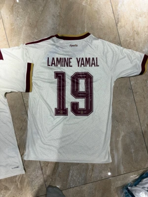 Lamine yamal  Spanien fotbollströja nr 19 - Säljer en vit Spanien fotbollströja från Adidas med Lamine Yamal och nummer 19 på ryggen. Tröjan har vinröda detaljer, gula inslag vid kragen och ärmslut samt officiellt landslagsemblem på bröstet. Tillverkad i lätt funktionsmaterial.