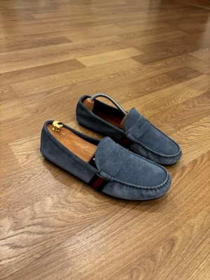 Gucci blå mocka loafers med randdetalj - Snygga loafers från Gucci i blå mocka med klassisk form och dekorativ rand i rött och mörkblått på sidan. Skorna har en stilren design med sömmar runt tån och en låg sula. Perfekta för dig som gillar en clean och avslappnad look. Nypris ca 8500 kr