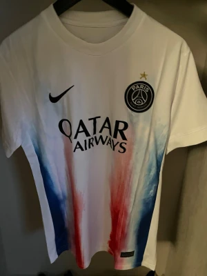 PSG matchtröja vit röd blå - Säljer en vit PSG matchtröja från Nike med tryckta detaljer i rött och blått framtill och baktill. Tröjan har Paris Saint-Germain-logga, Qatar Airways sponsortryck och en guldstjärna ovanför klubbmärket. Materialet är lätt och andas, perfekt för fotboll eller träning.