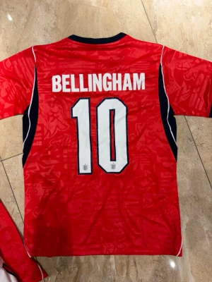 England borta  Bellingham #10 -S - Säljer en röd England fotbollströja från Nike med Bellingham och nummer 10 på ryggen. Tröjan har svarta och vita detaljer, korta ärmar och England-emblemet på bröstet. Perfekt för dig som vill stötta landslaget på match eller träning.