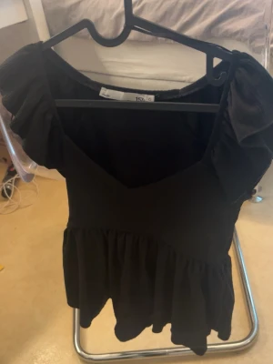Svart kort klänning från 157 - Säljer en svart kort klänning från 157 med volangärmar och v-ringad hals. Klänningen har en lös passform och volang nedtill som ger en söt och trendig look. Perfekt för dig som gillar stilrena plagg med en twist.