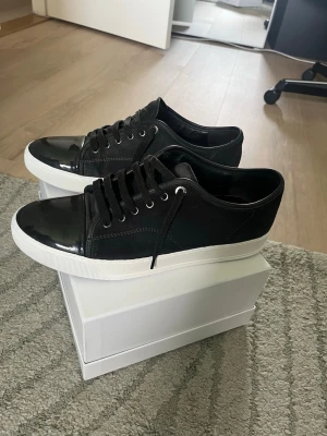 Svarta sneakers med lackdetalj lanvin - Stilrena svarta sneakers med vit sula och snygg lackad tå. Ovandelen är i mockaliknande material och skorna har svarta skosnören samt metallöljetter. Perfekta för dig som vill ha en clean och modern look.