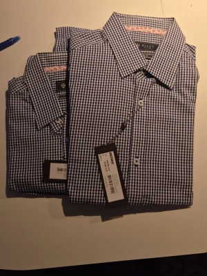 Blårutig skjorta från Riley - Säljer en klassisk blårutig skjorta från Riley i slim fit-modell. Skjortan har vit-blått rutmönster, knappar framtill och krage. Perfekt för dig som gillar stilrena och tidlösa plagg. 200kr st
