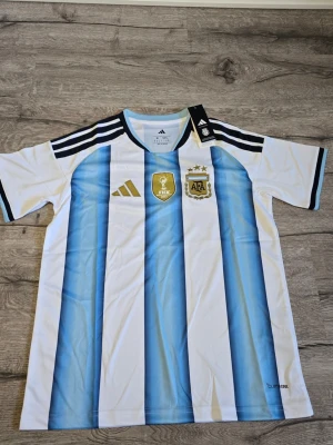 Argentina fotbollströja Adidas - Säljer en officiell Argentina fotbollströja från Adidas med klassiska blå och vita vertikala ränder. Tröjan har svarta detaljer på axlarna, FIFA- och AFA-emblem på bröstet samt rund hals. Perfekt för dig som älskar fotboll och vill stötta ditt lag.